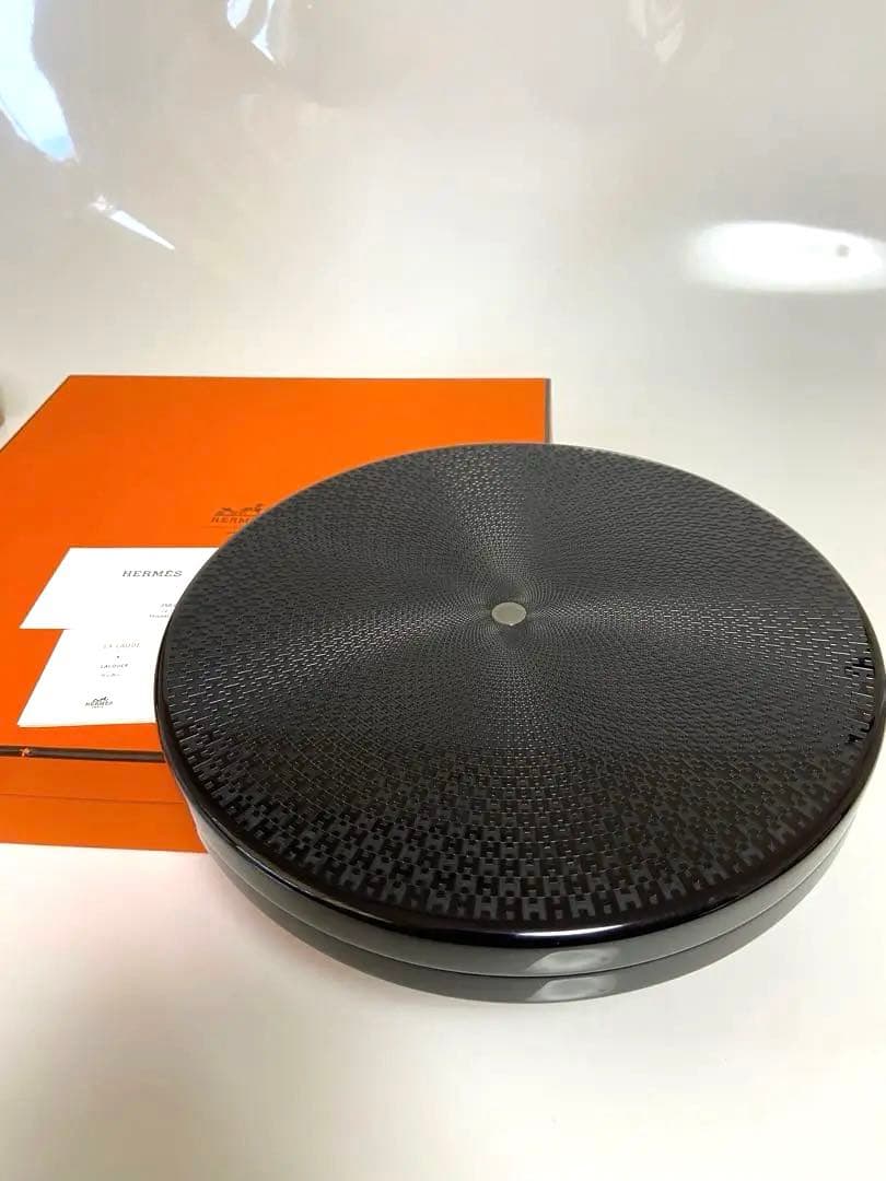 Round dark lacquer wooden box with a textured radial pattern and small central metal dot, shown with an orange Hermès box and papers behind it.