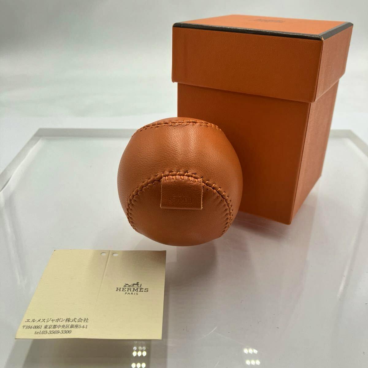 Orange leather baseball ornament displayed with its orange Hermès box and a Hermès card on a clear surface.