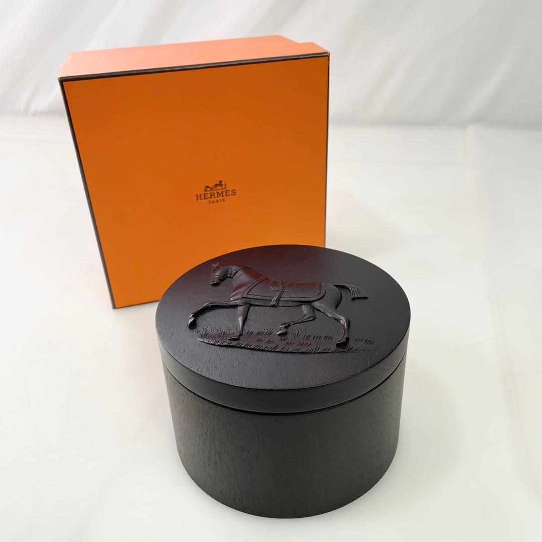 Round mahogany wooden Hermès Twilly box with a carved horse motif on the lid, shown in front of an orange Hermès box.