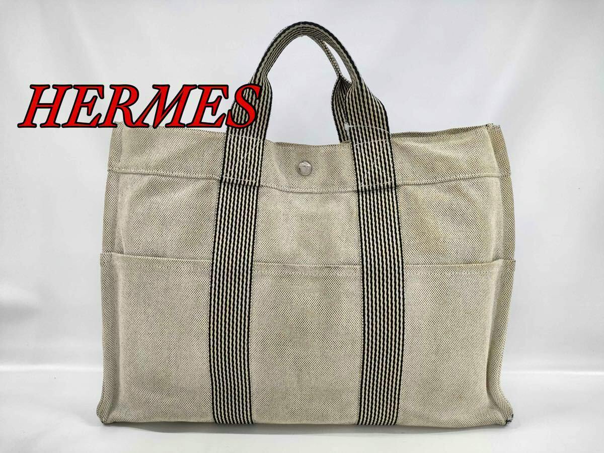 Front view of an ivory and black Hermès canvas tote bag with striped handles and exterior pockets against a white background.
