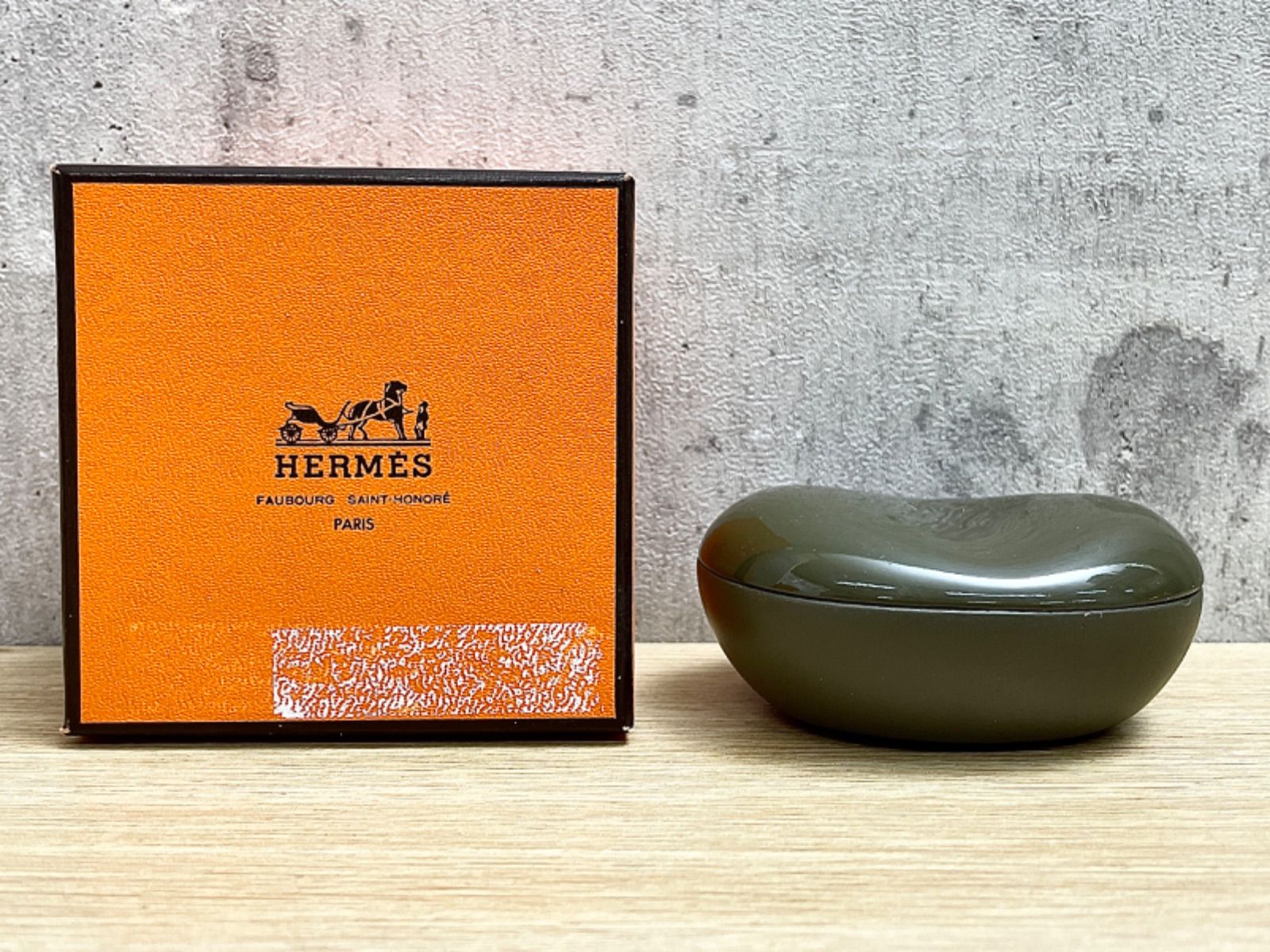 Olive wooden bean-shaped small case displayed next to an orange Hermès box on a wooden surface.