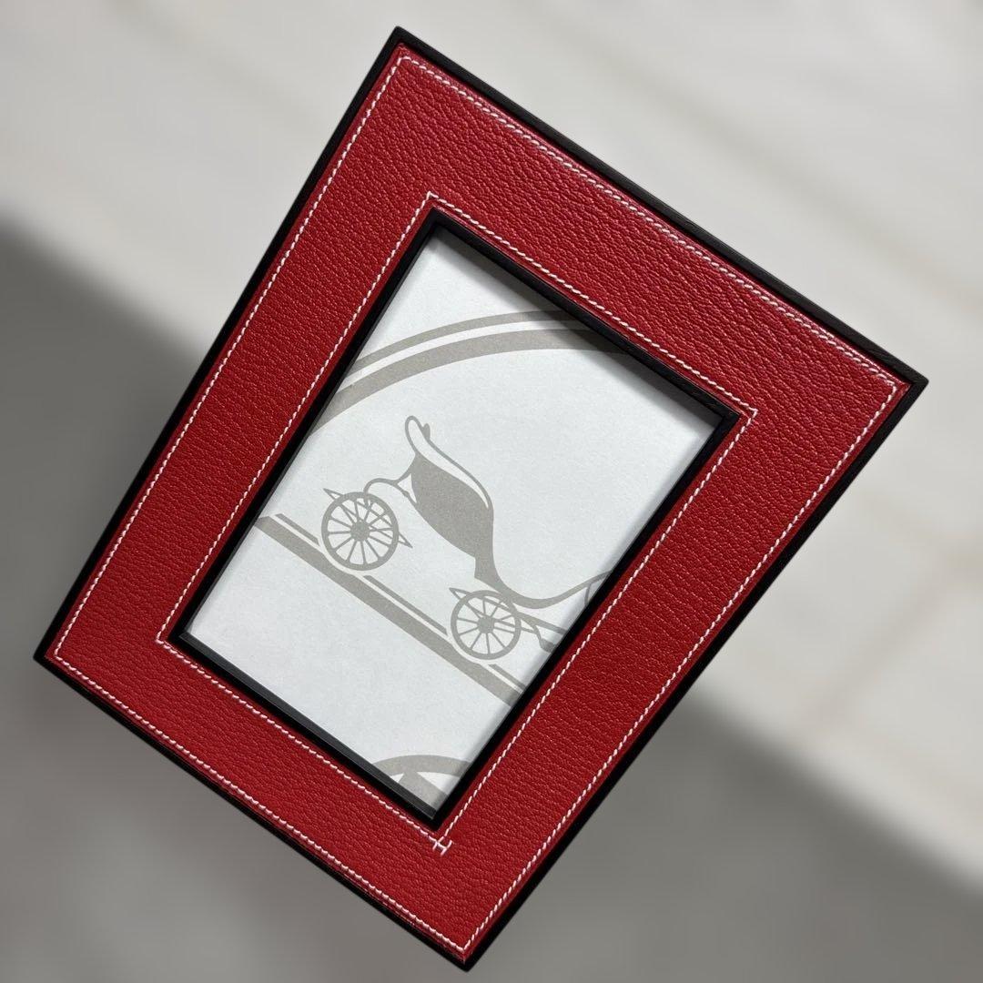 Red Hermès photo frame with white stitching and a paper insert showing a carriage motif.