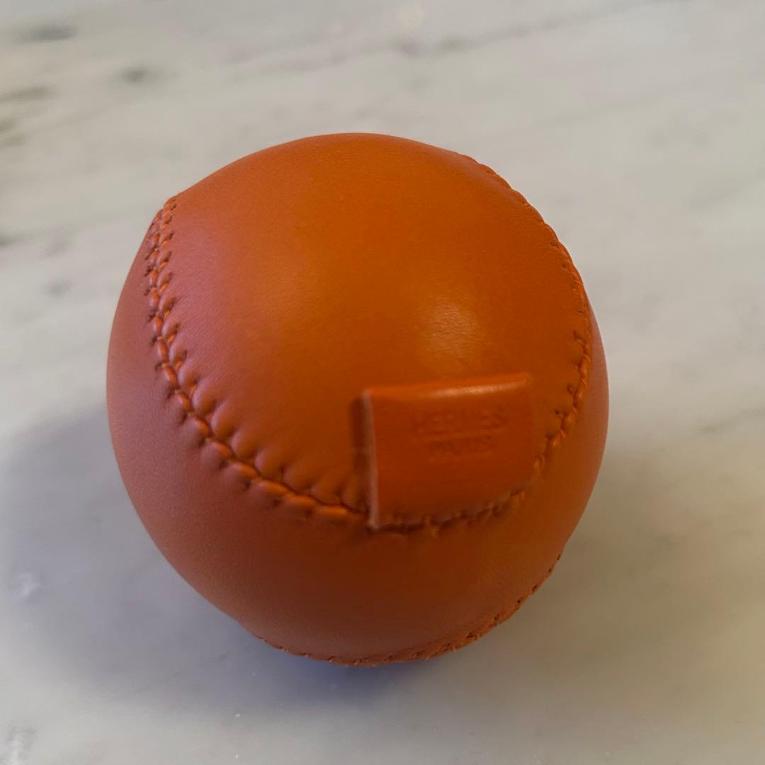 Close-up of an orange Hermès leather baseball showing stitching and a small stamped tab.