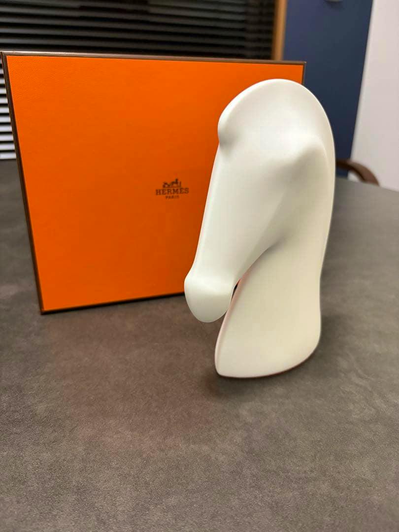 White ceramic horse-head Hermès paperweight displayed in front of an orange Hermès box on a dark surface.