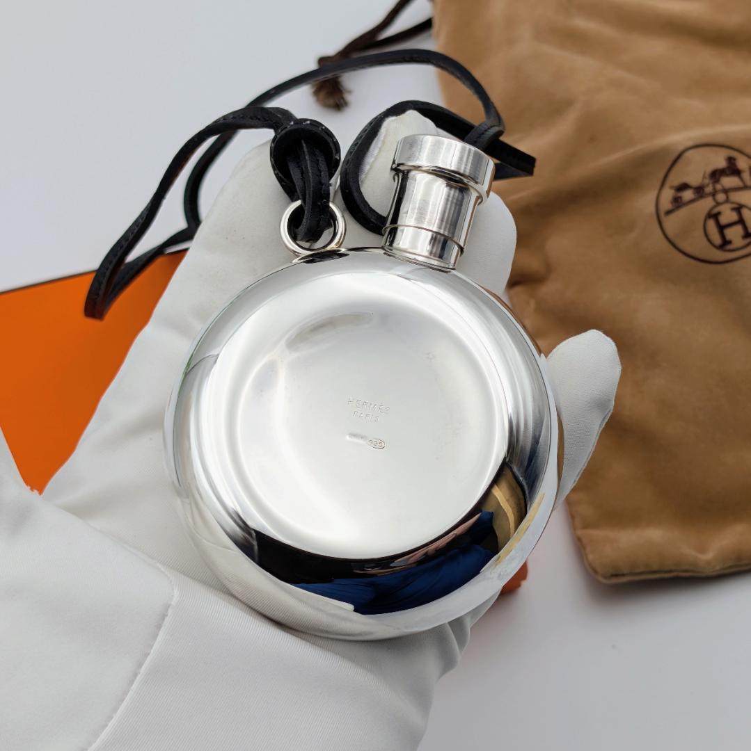 Round silver (Silver 925) Hermes flask with screw cap and leather strap held in a white gloved hand with a Hermes dust bag partially visible.