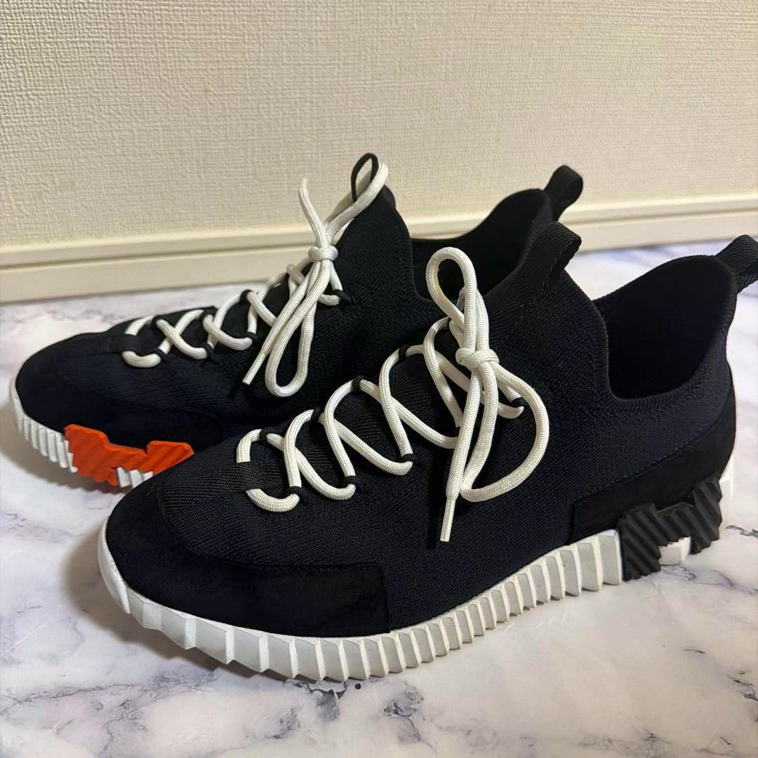 Pair of black textile Hermès sneakers with white laces and a ridged white sole with an orange accent, shown on a marble-like surface.