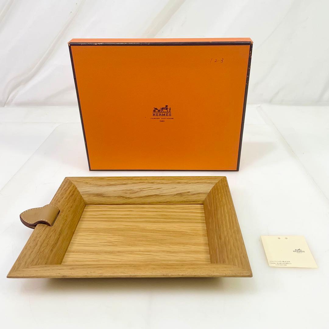 Wooden Hermès Vid Poche atrium tray displayed on a white surface with its orange Hermès box and a small tag behind it.