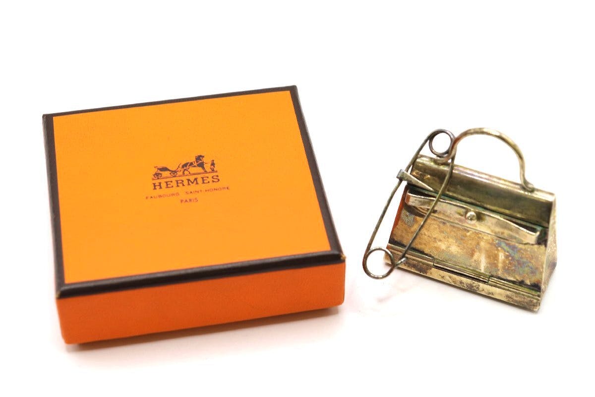 Silver HERMES Kelly bag–shaped pill case charm placed next to an orange HERMES box.