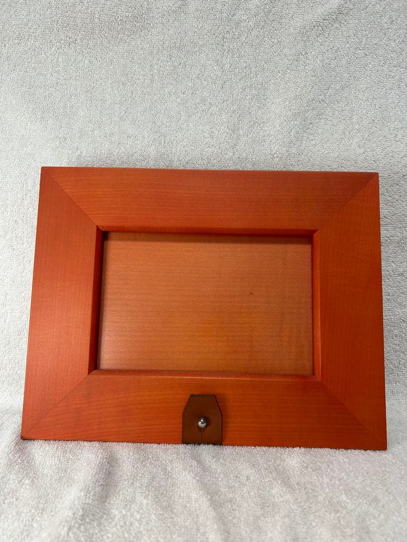 Front view of an orange wooden photo frame with a small brown tab at the bottom, placed on a light fabric background.