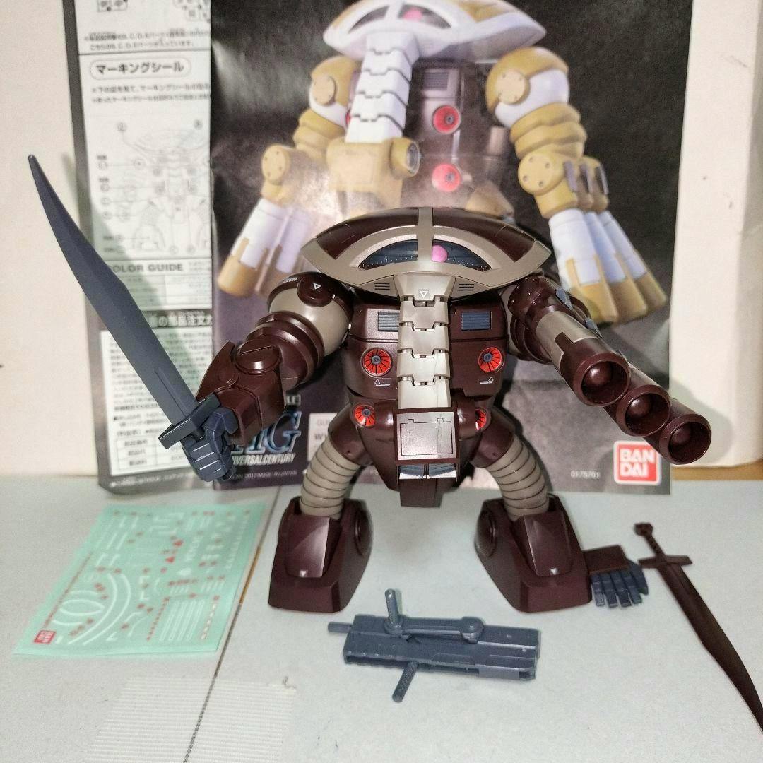 Front view of the assembled HG 1/144 Juagg (Gundam model) plastic figure in brown with a sword and accessories, with the product box and manual visible behind it.