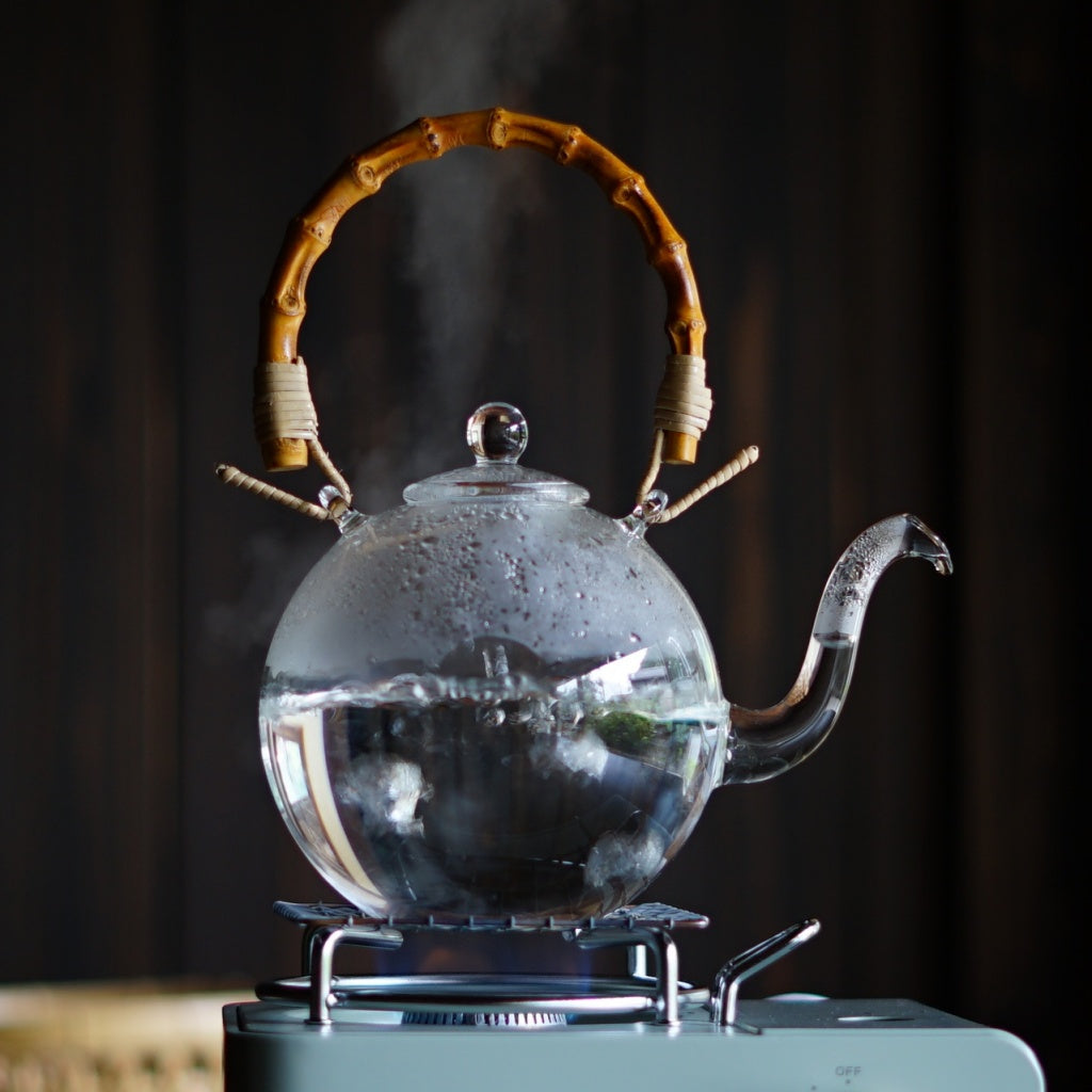 Clear glass ball pot with a bamboo handle boiling on a stovetop.