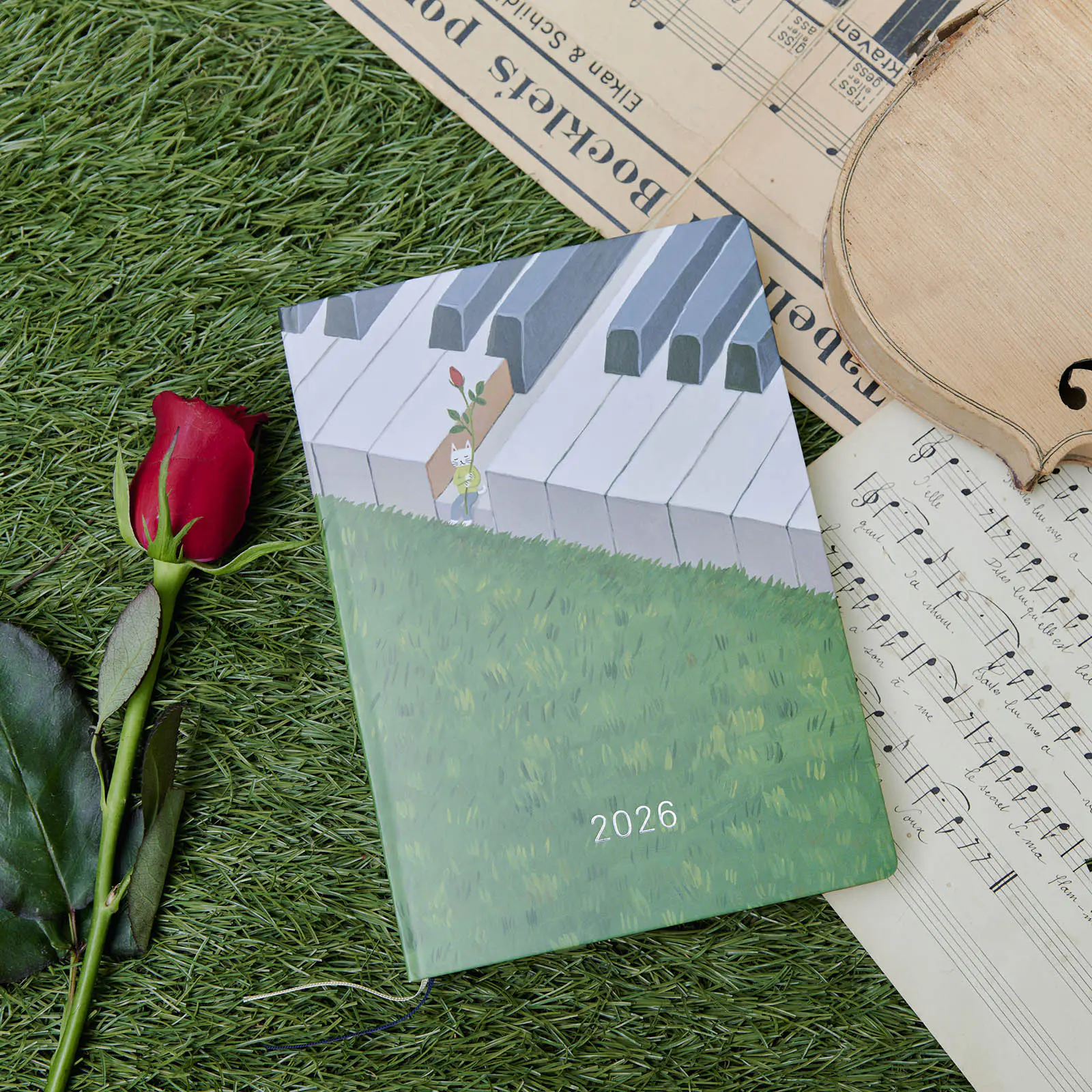Main photo of the Hiroko Kubota: Waiting on the Chord HON ENG edition 2026 diary with a piano-key illustrated cover placed on grass beside a red rose and sheet music.