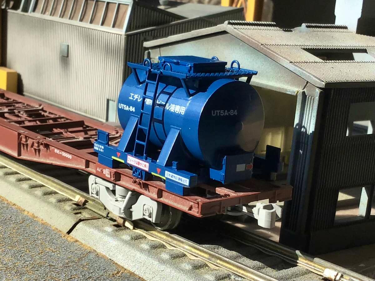 Blue HO-scale UT5A-84 tank container model mounted on a flat railcar in a diorama setting.