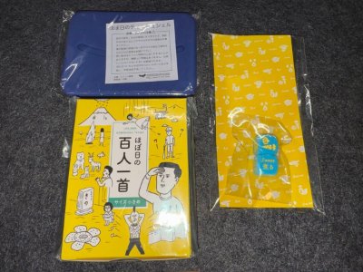 Three items of the Hobonichi goods set on a gray surface: a blue tissue pouch, a yellow Hyakunin Isshu booklet, and a packaged dice on yellow backing.
