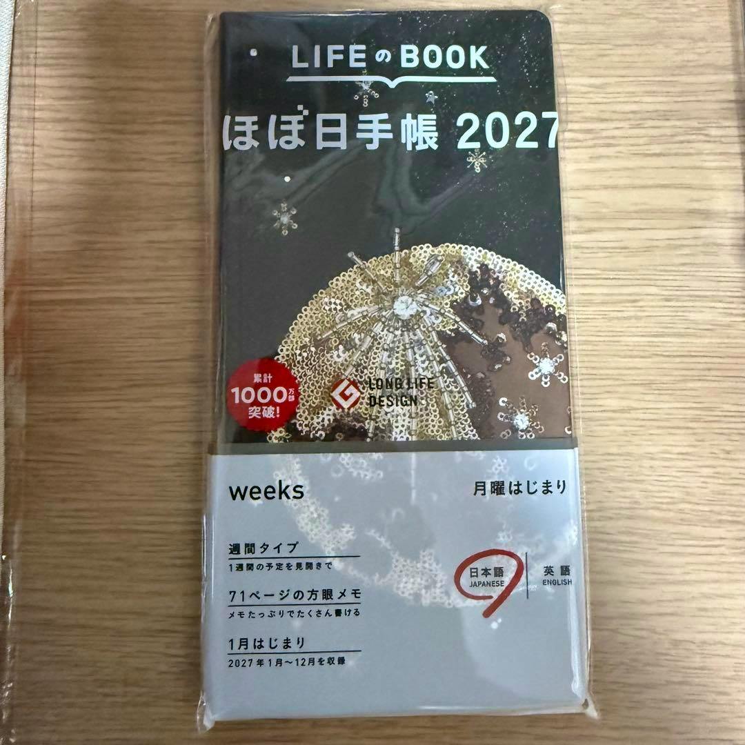 Front view of the ほぼ日手帳 2027 weeks diary (Starlight tanakadaisuke) in its plastic sleeve showing the cover and white paper band.