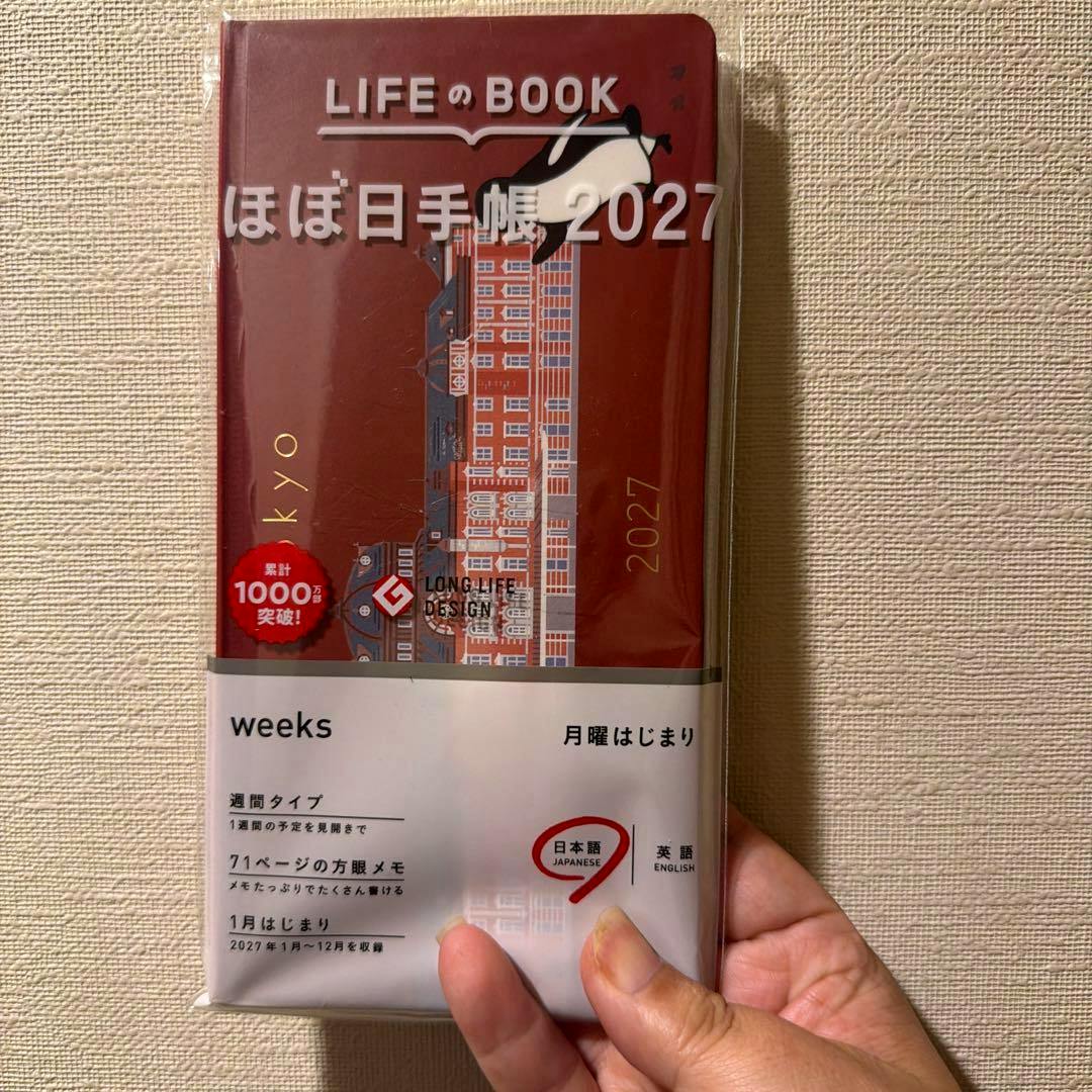 Hand holding the front of the Hobonichi 2027 weeks diary in its plastic sleeve showing the red Tokyo Station Suica Penguin cover.