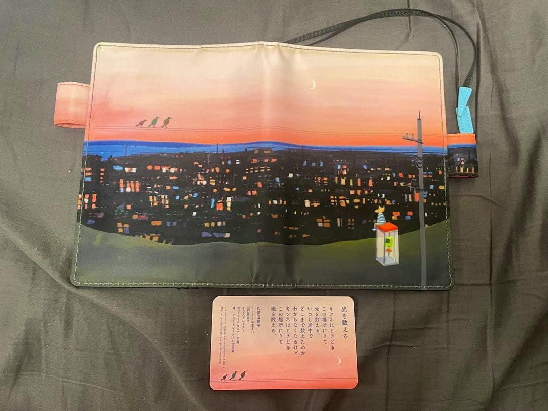 An open ほぼ日手帳 カバー 久保田寛子 光を数える A6 cover showing the illustrated cityscape sunset exterior design with a small informational card placed below.
