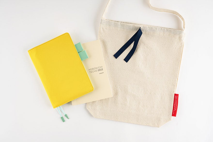 Hobonichi Techo reusable tote bag with shoulder strap and navy ribbon tab shown next to two Hobonichi Techo notebooks.