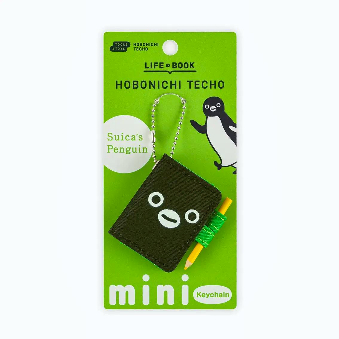 A Hobonichi Techo “Suica’s Penguin” mini keychain with a small pencil attached, displayed on a green product backing card.