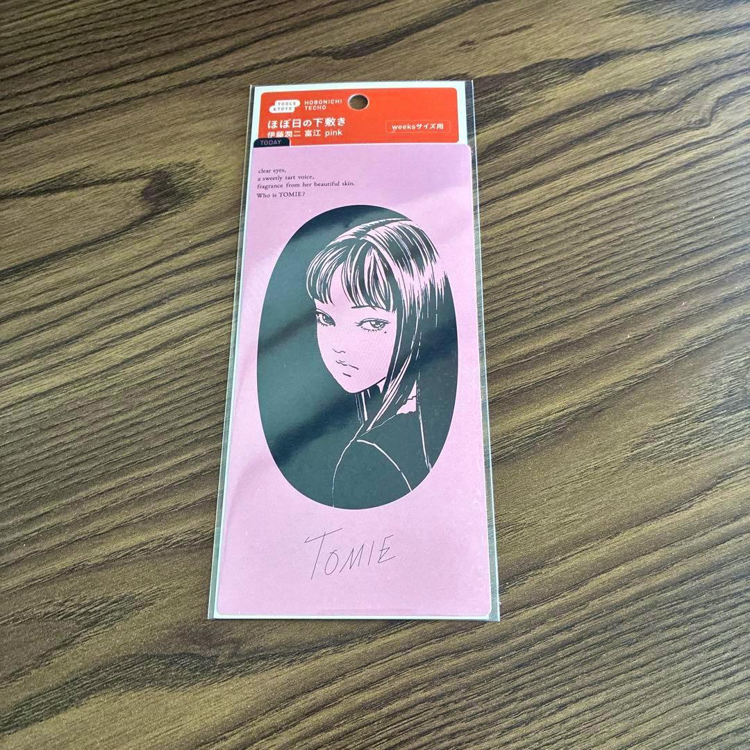 Packaged pink Hobonichi weeks-size acrylic board featuring Junji Ito’s "Tomie" illustration on a wooden surface.