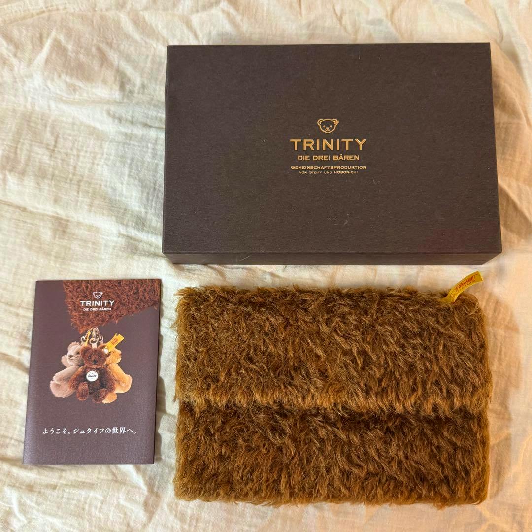 A folded chocolate-colored furry Weeks wallet (Steiff) displayed next to a TRINITY box and a booklet.