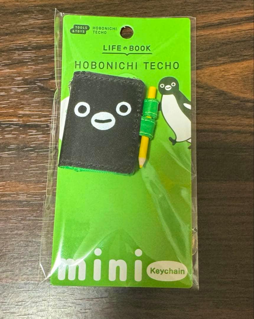 Packaged Hobonichi Techo Suica mini keychain showing a small black notebook-shaped charm and a yellow pencil on a green backing card in plastic packaging.