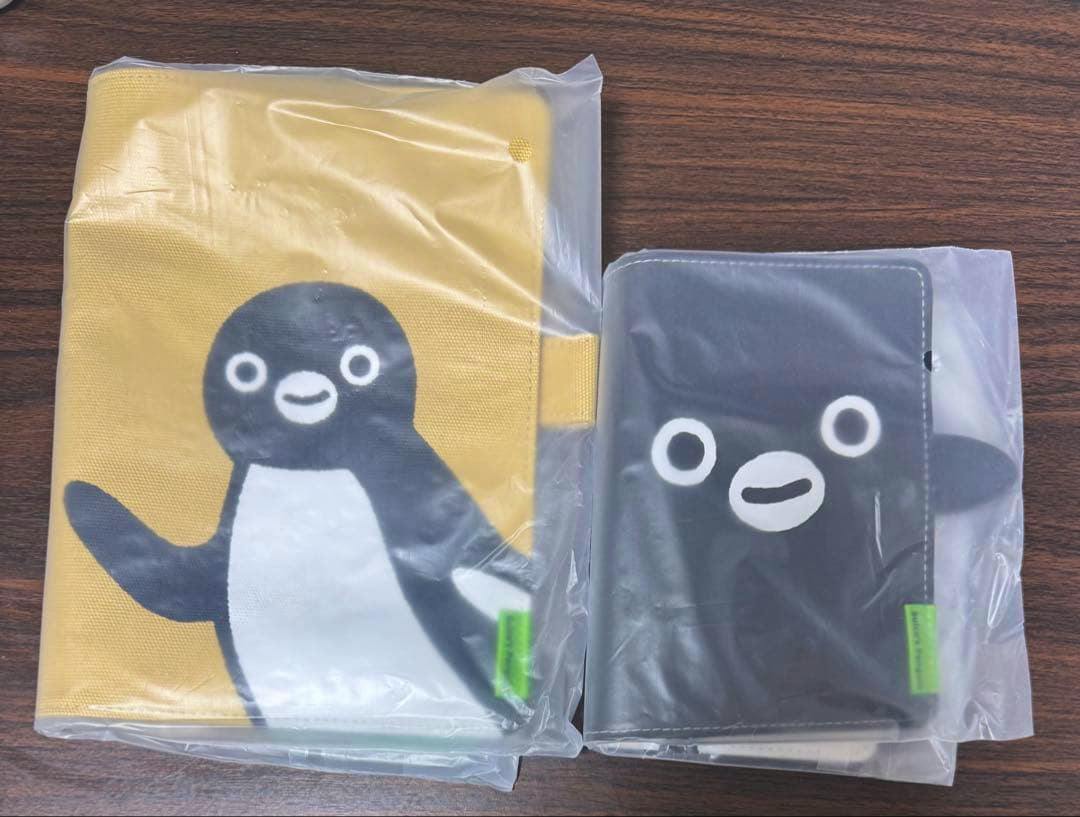 Two Suica-themed Hobonichi notebook covers in plastic packaging, a yellow penguin design and a black penguin design, shown side by side on a wooden surface.