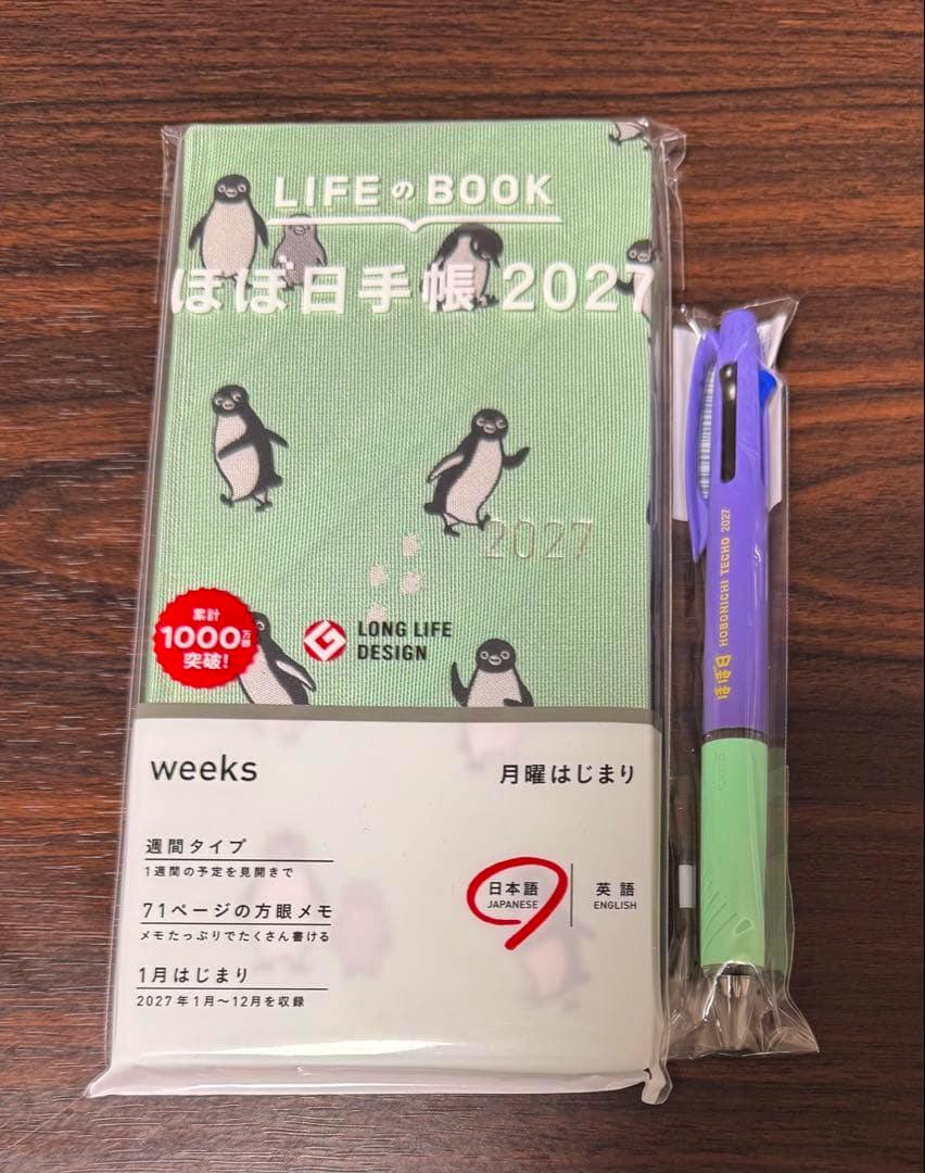 Packaged ほぼ日手帳 Suica weeks 2027 diary with a penguin-pattern cover and a packaged ballpoint pen on a wooden surface.