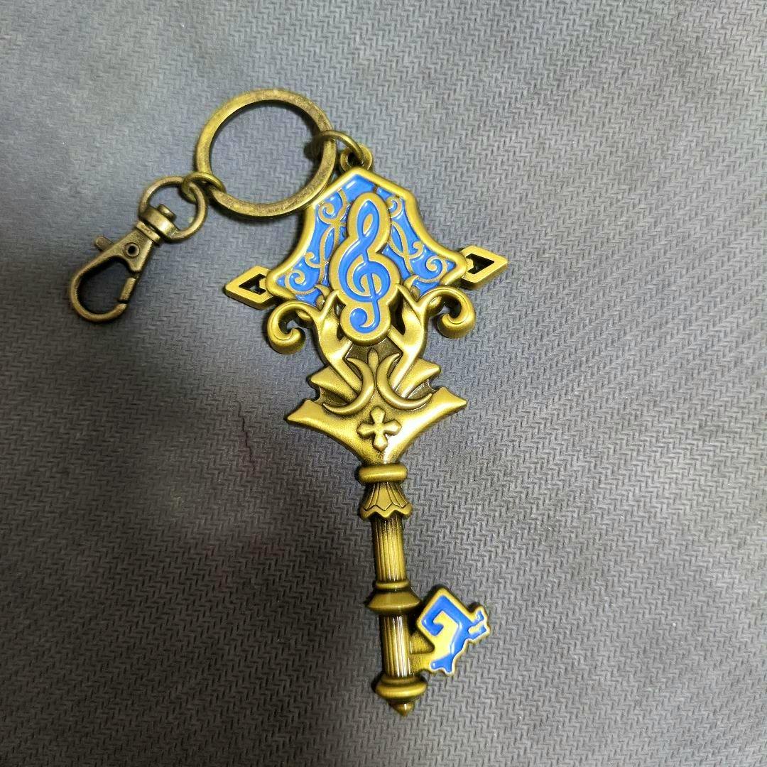 Front view of the ホロライブ Advent 1周年記念 モチーフキーホルダー ネリッサ keyholder showing a gold-tone ornate key-shaped charm with a blue treble-clef motif, metal ring, and clasp.