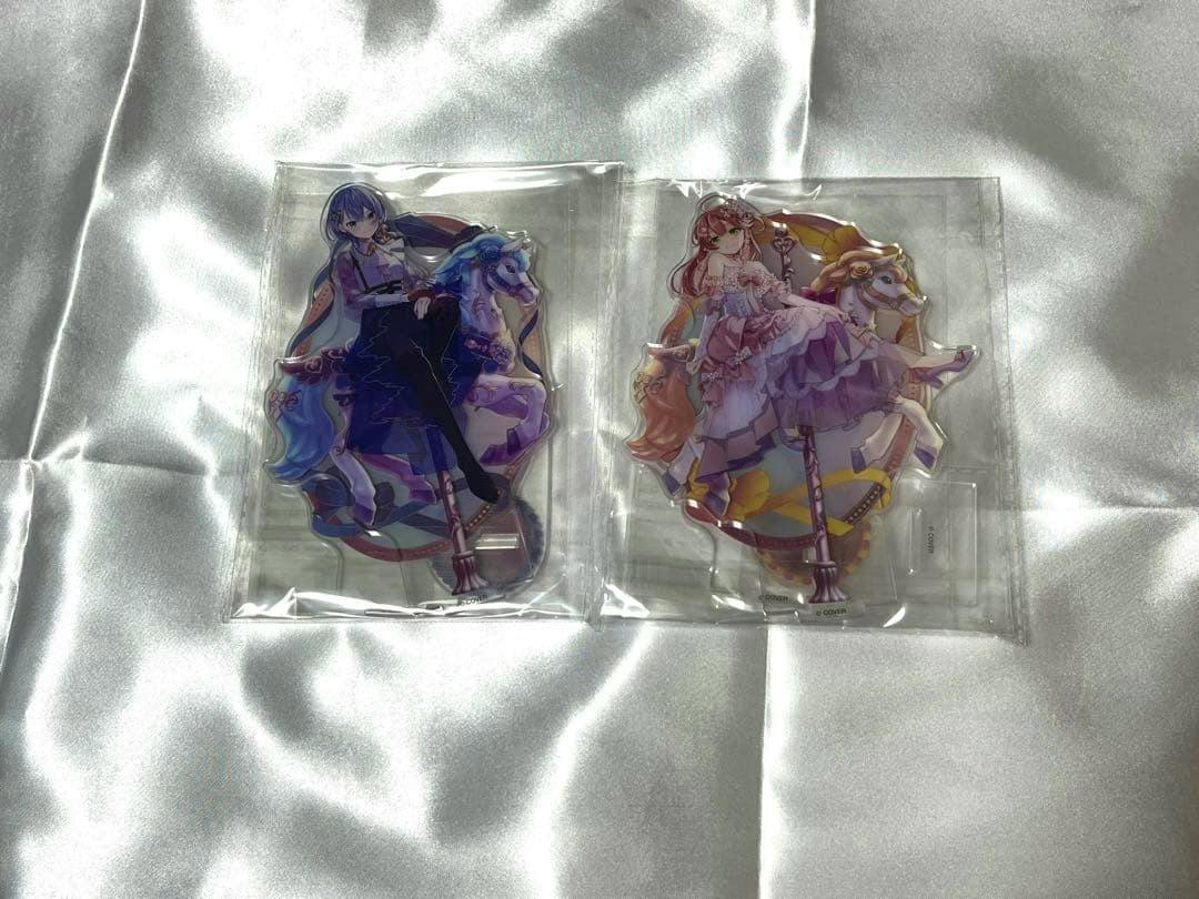 Two packaged acrylic character stands featuring Hololive characters Hoshimachi Suisei and Sakura Miko laid on a silver fabric background.