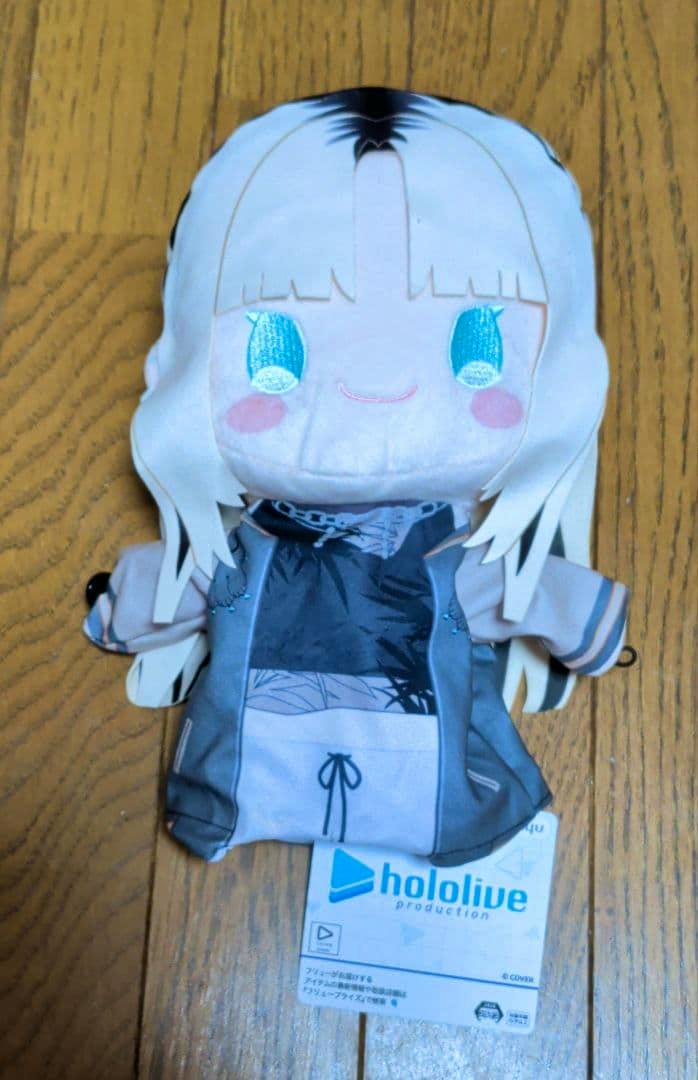 Front view of the Hololive puppet plush toy showing the character's face, clothing, and attached Hololive tag on a wooden floor.