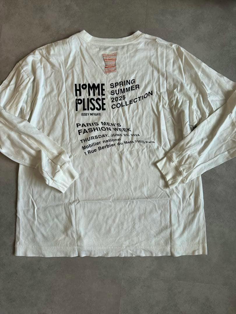 White long-sleeve cotton T-shirt laid flat showing the back with "HOMME PLISSÉ ISSEY MIYAKE" printed text and event details.