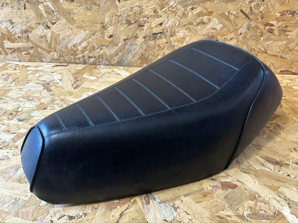 Black motorbike seat placed on a plywood background, shown at a front-side angle.