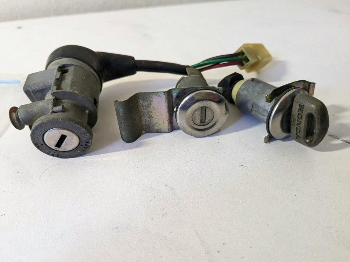 Three motorbike ignition lock components including an ignition key switch with attached wiring and two lock cylinders on a white background.