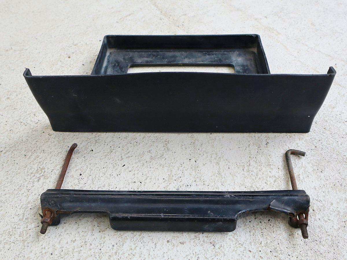 A Honda TN360 car battery stay showing a black battery tray and a retaining bar with rusted rods on a concrete surface.