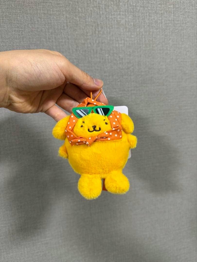 Yellow Pom Pom Purin plush mascot wearing an orange polka-dot headscarf and green sunglasses, held by a hand.