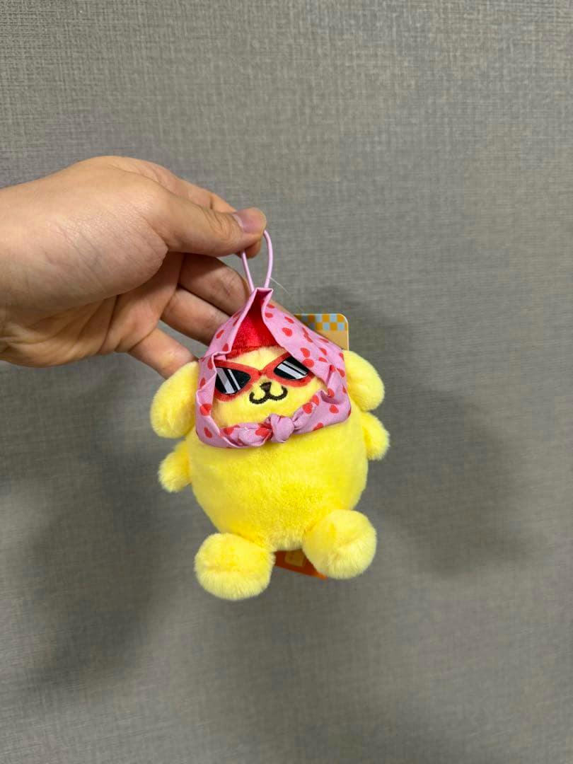 Front view of a yellow Pompompurin plush mascot wearing a pink hood and red sunglasses, held by a hand.
