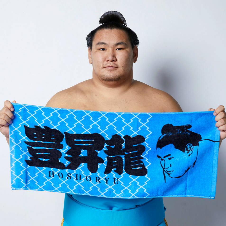 A sumo wrestler holds a blue Hoshoryu face towel showing large Japanese characters and a head illustration.