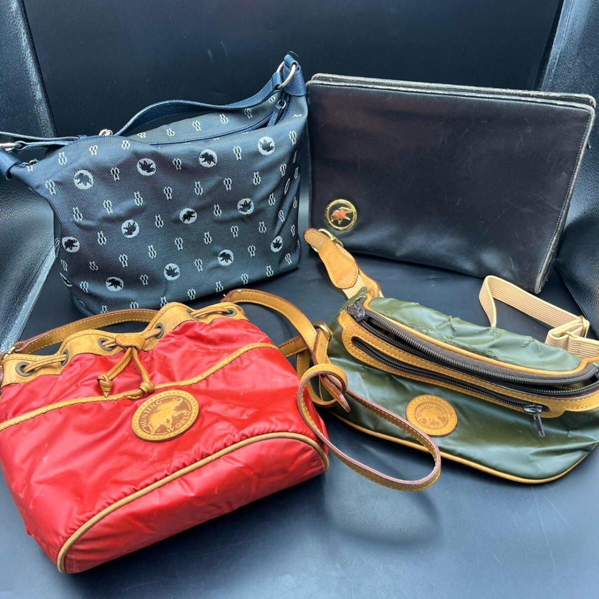 Four Hunting World bags (navy patterned shoulder, black clutch, red drawstring handbag, and green waist pouch) arranged together on a dark surface.