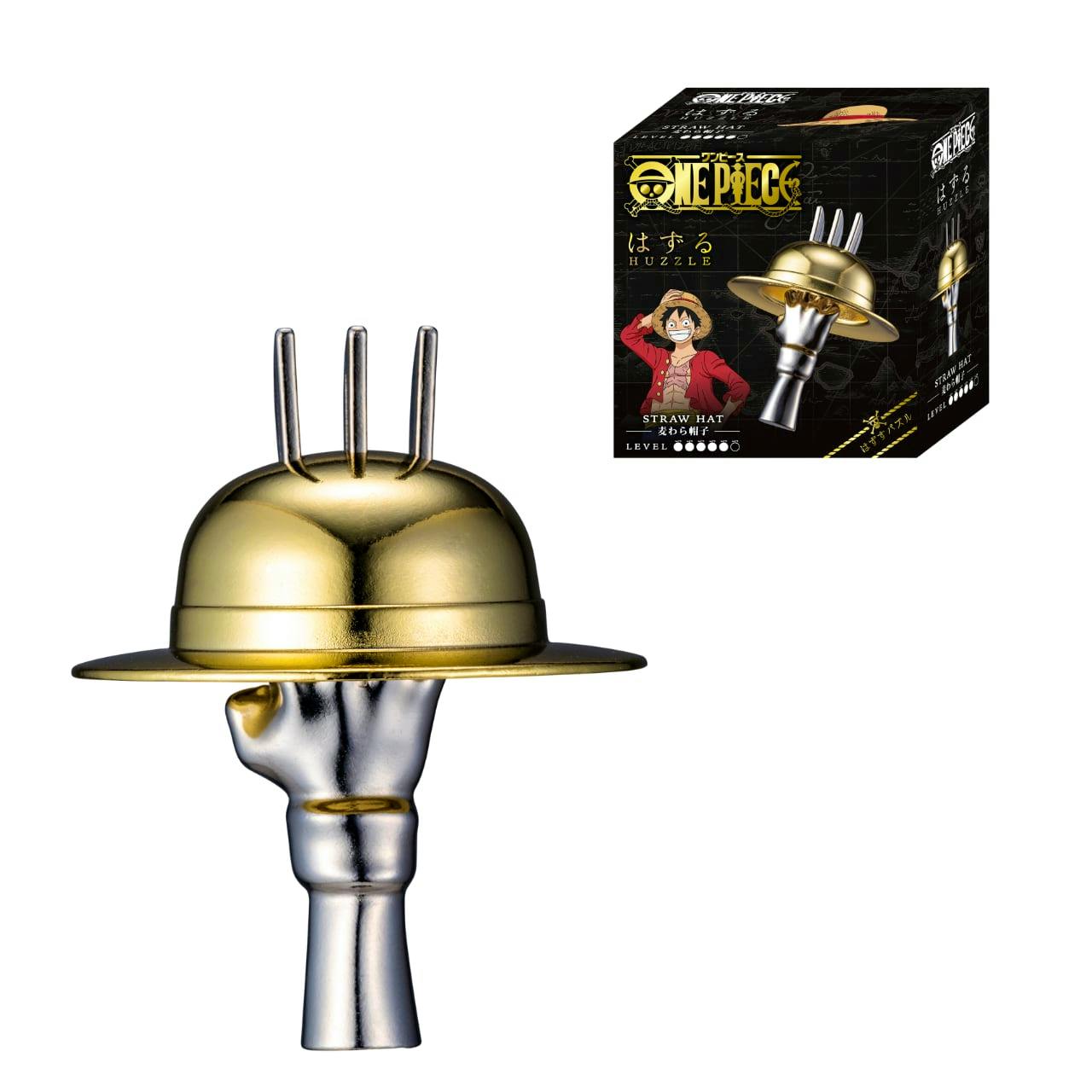 A metal puzzle shaped like a straw hat held on a silver metallic hand, with the product box showing ONE PIECE branding visible in the corner.