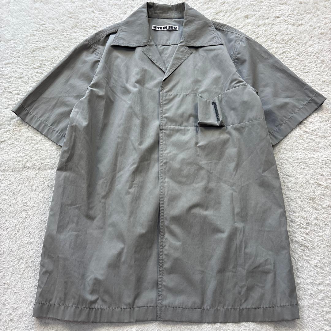 Front view of a grey polyester HYEIN SEO men's smoker's shirt with short sleeves and a chest pocket, laid flat on a light textured surface.