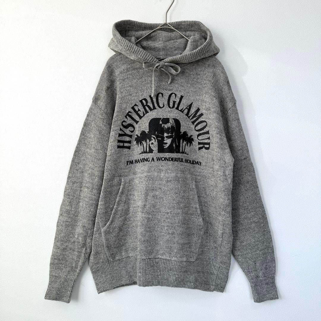 Gray linen-mix knit hoodie (sweater) with a black HYSTERIC GLAMOUR chest graphic and front kangaroo pocket, hanging on a hanger against a white background.