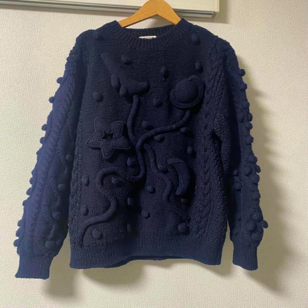 Navy 3D knit wool sweater with raised planet and star motifs hung on a wooden hanger against a wall.