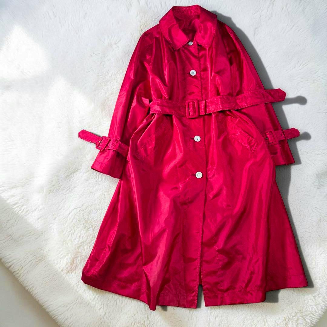 A long red men's trench coat laid flat on a white surface, showing buttons and a belt.