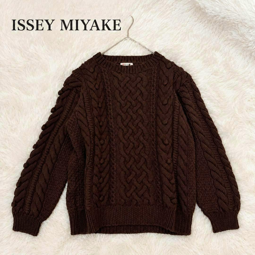 Brown cable-knit wool sweater on a hanger laid flat on a white furry background.