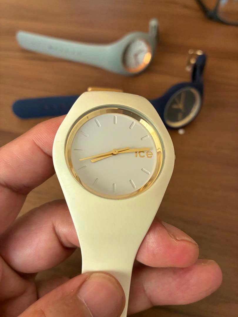 Close-up of a white ICE WATCH wristwatch with gold accents held in a hand, with two other watches blurred on a wooden surface in the background.