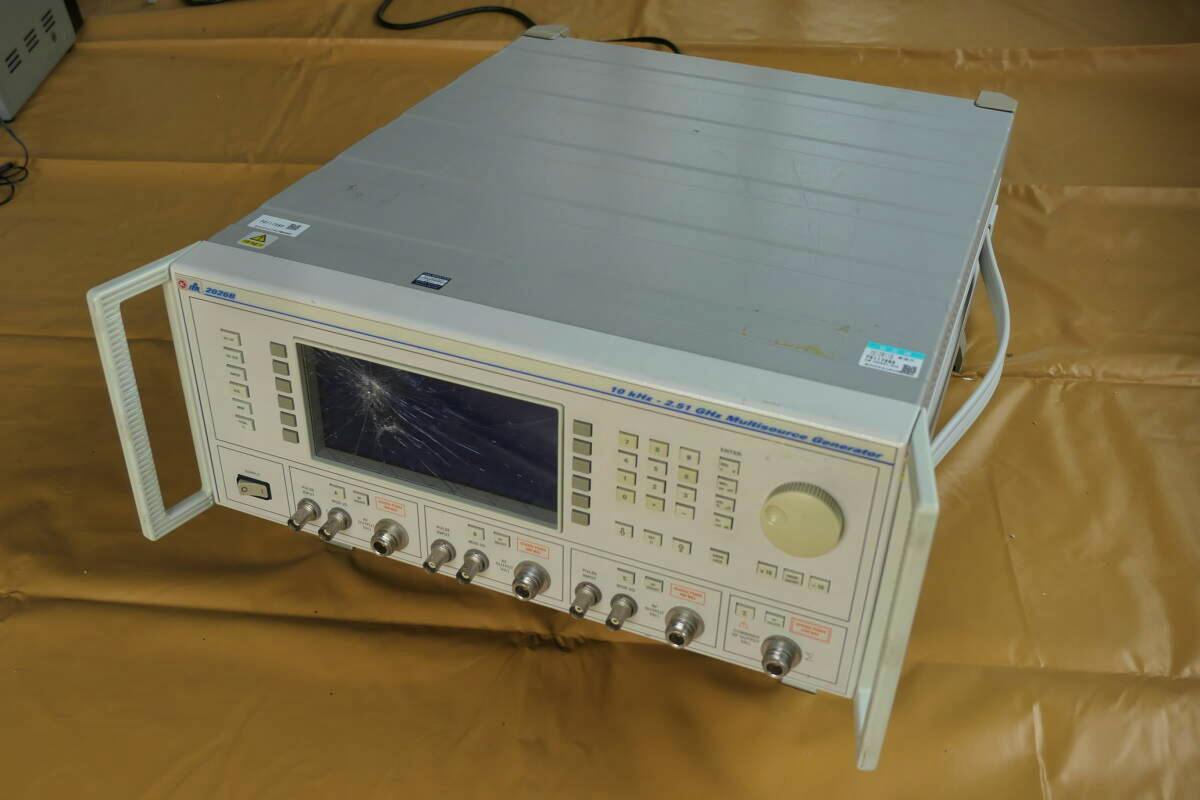 Front and top view of the ITL 2026B multisource signal generator showing its front panel, connectors, and a cracked display.
