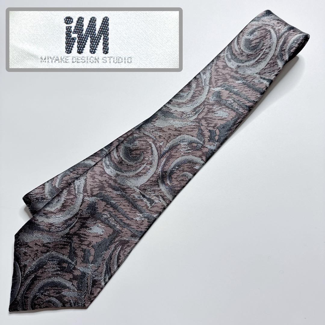 Silver patterned silk necktie laid flat on a white background with an inset showing the im ISSEI MIYAKE label.