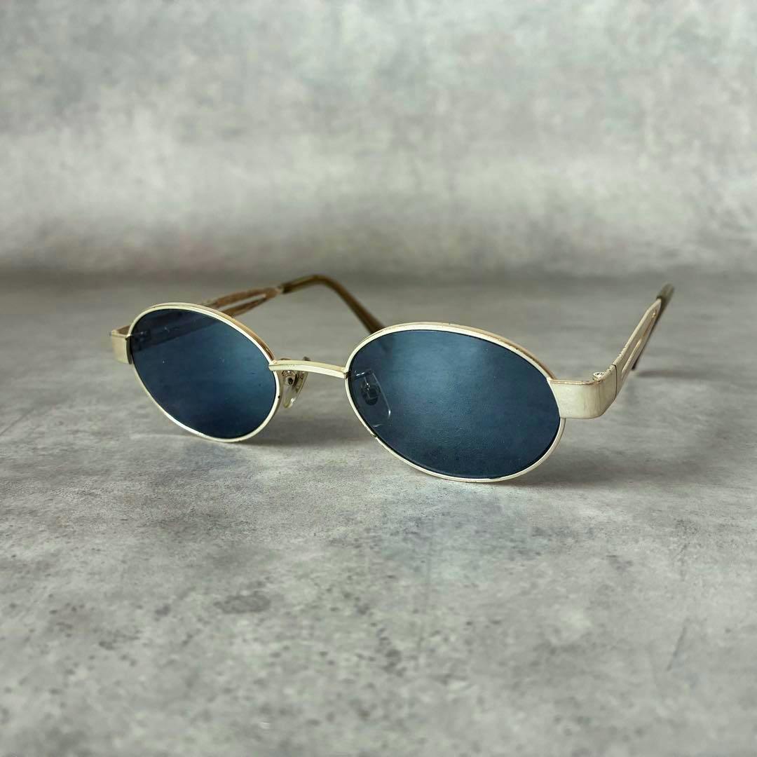A pair of IM ISSEY MIYAKE sunglasses with blue oval lenses and gold-tone frames on a gray surface.