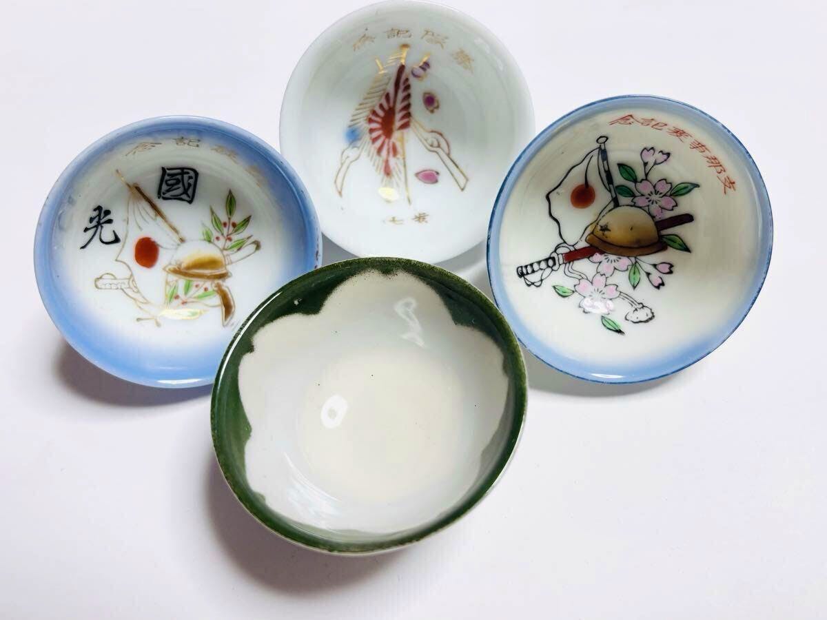 Four small ceramic military sake cups (bowls) on a white background, three showing painted military-themed interior designs and one with a green rim.